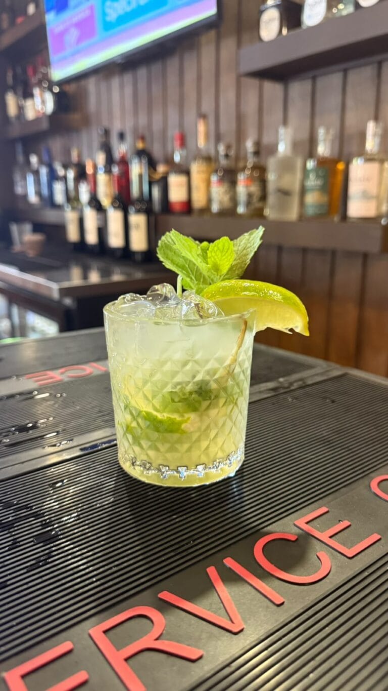 A refreshing mojito cocktail garnished with a lime wedge and mint leaves, placed on a bar counter with a blurred background of various liquor bottles in a bar on Roatan, Honduras.