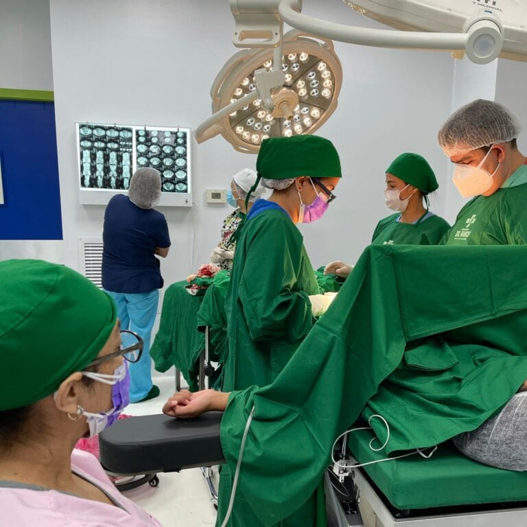 Surgeons and medical staff performing surgery in an operating room in Roatan, Honduras, with surgical lights overhead and X-ray images displayed on a screen in the background.