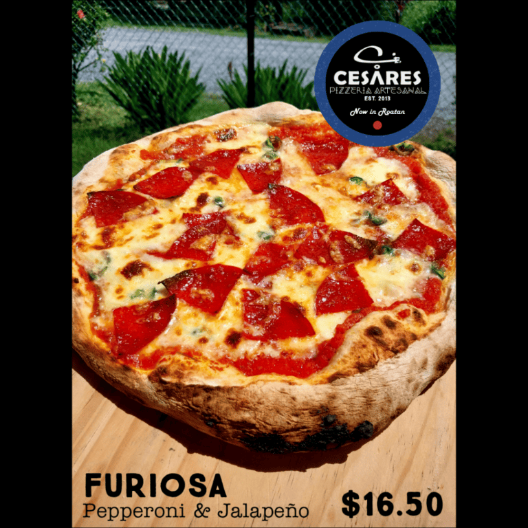 A pizza with pepperoni and jalapeños sits on a wooden table outdoors in Roatan, Bay Islands, Honduras, with greenery and a chain-link fence in the background.