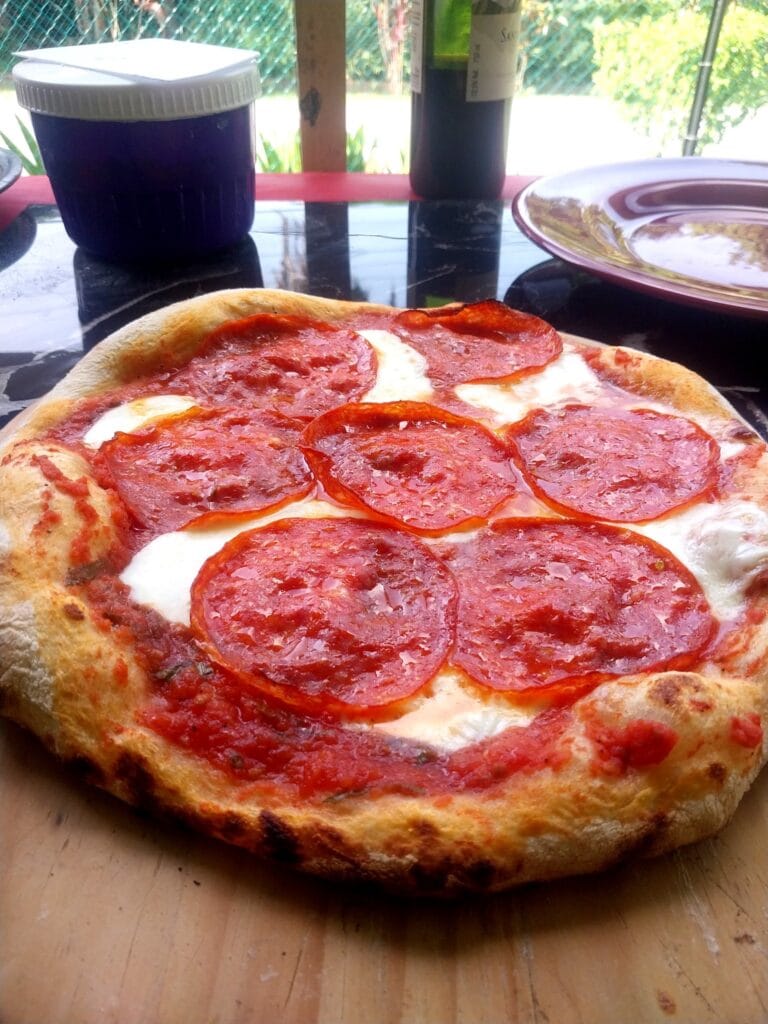 A freshly baked pepperoni pizza with melted cheese and crispy crust on a wooden surface in Roatan, Honduras, with colorful plates and bottles in the background.