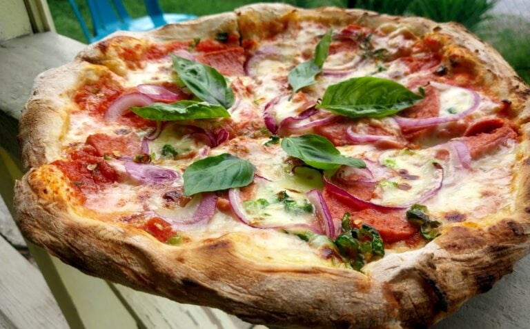 Freshly baked pizza topped with basil leaves, red onions, tomato sauce, and melted cheese, sitting on a wooden surface outdoors in Roatan, Honduras.