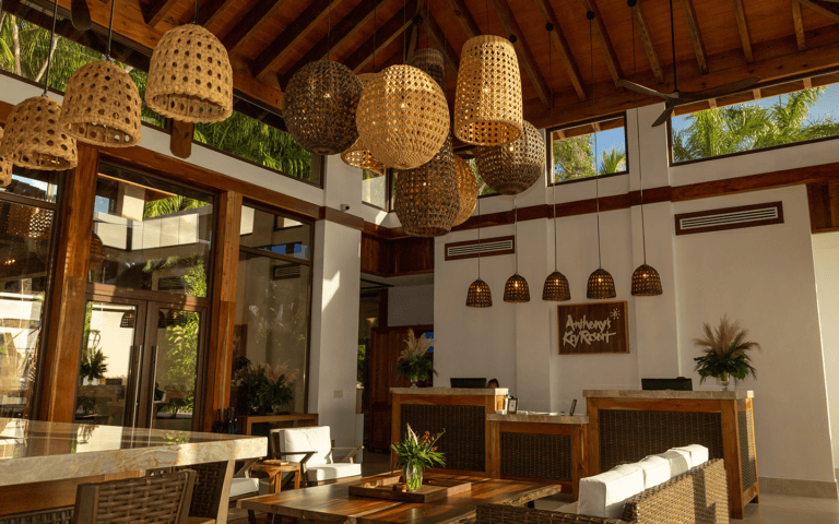 The interior of a tropical restaurant in Roatan, Honduras, featuring a high wooden ceiling with woven pendant lights and large windows revealing lush greenery outside.