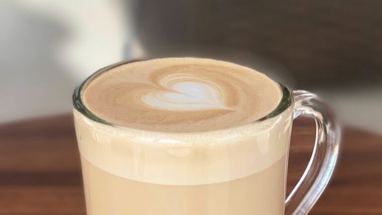 A close-up of a glass mug filled with a creamy latte topped with latte art resembling a heart or flower design, set on a wooden surface in Roatan, Honduras.