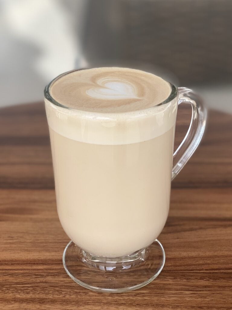 A clear glass mug filled with a creamy latte featuring heart-shaped latte art on the surface, placed on a wooden table with a blurred background.