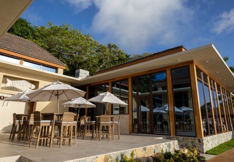 Modern restaurant with large glass windows and a wooden exterior, surrounded by patio tables with white umbrellas, under a clear blue sky in Roatan, Honduras.