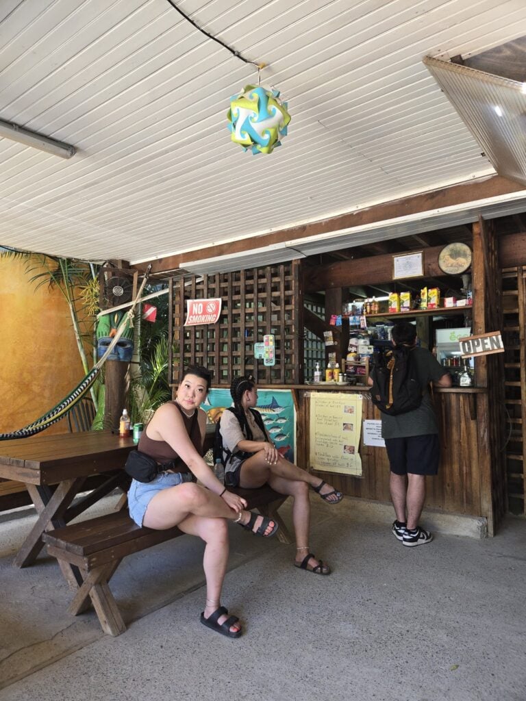 Indoor seating area at a restaurant or cafe in Roatan, Bay Islands, Honduras, with three people sitting at a wooden table and a counter behind it where a staff member is preparing orders, featuring tropical decor including a carved mask.