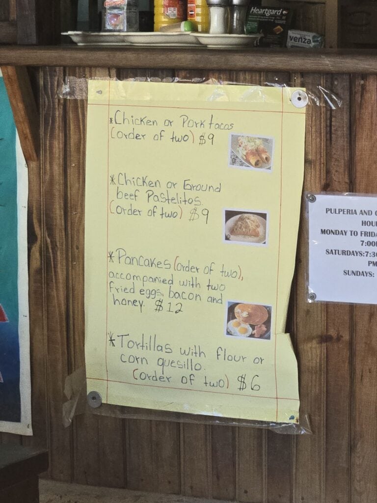 A handwritten menu on yellow paper listing food options like chicken or pork tacos, beef pasteles, and tortillas with flour or corn quesillo, attached to a wooden wall in Roatan, Honduras, with small photos of some dishes.