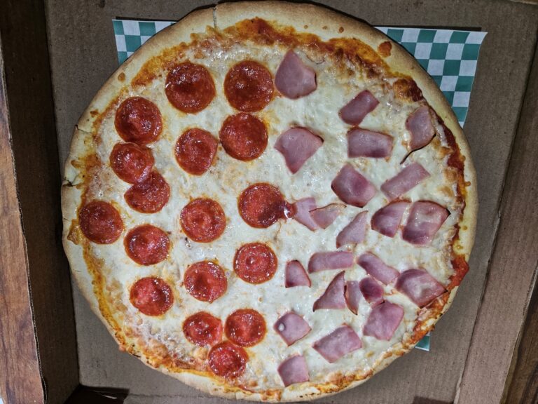 A large pizza with pepperoni slices on the left side and ham slices on the right side, placed inside a brown cardboard pizza box on a wooden surface in Roatan, Honduras.