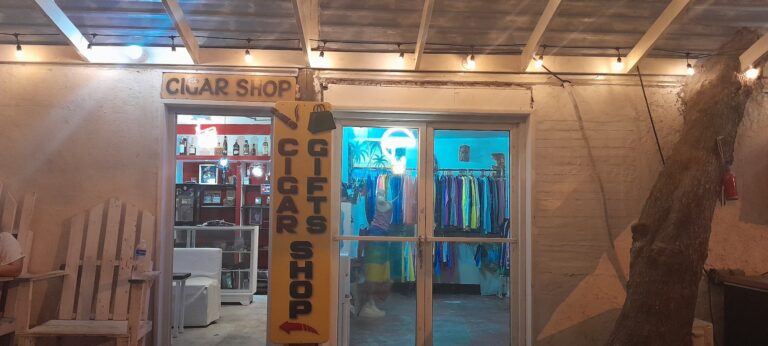 The entrance of a cigar shop in Roatan, Honduras, featuring a yellow sign with black lettering and a display window with colorful clothing and palm tree murals inside.