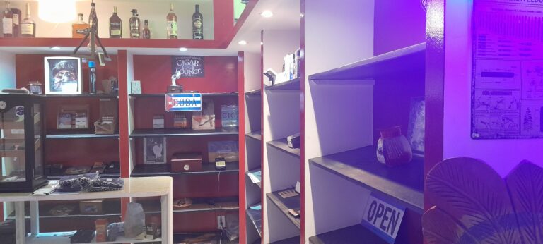 Indoor view of a convenience store or shop with shelves stocked with liquor bottles, a glass display case, and a sign indicating it is in Roatan, Honduras.