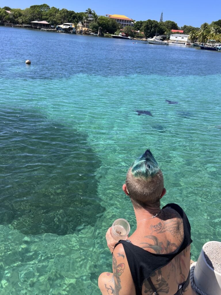 A tattooed person sits on a boat overlooking clear turquoise water, reef formations, and the shoreline of Roatan, Honduras.