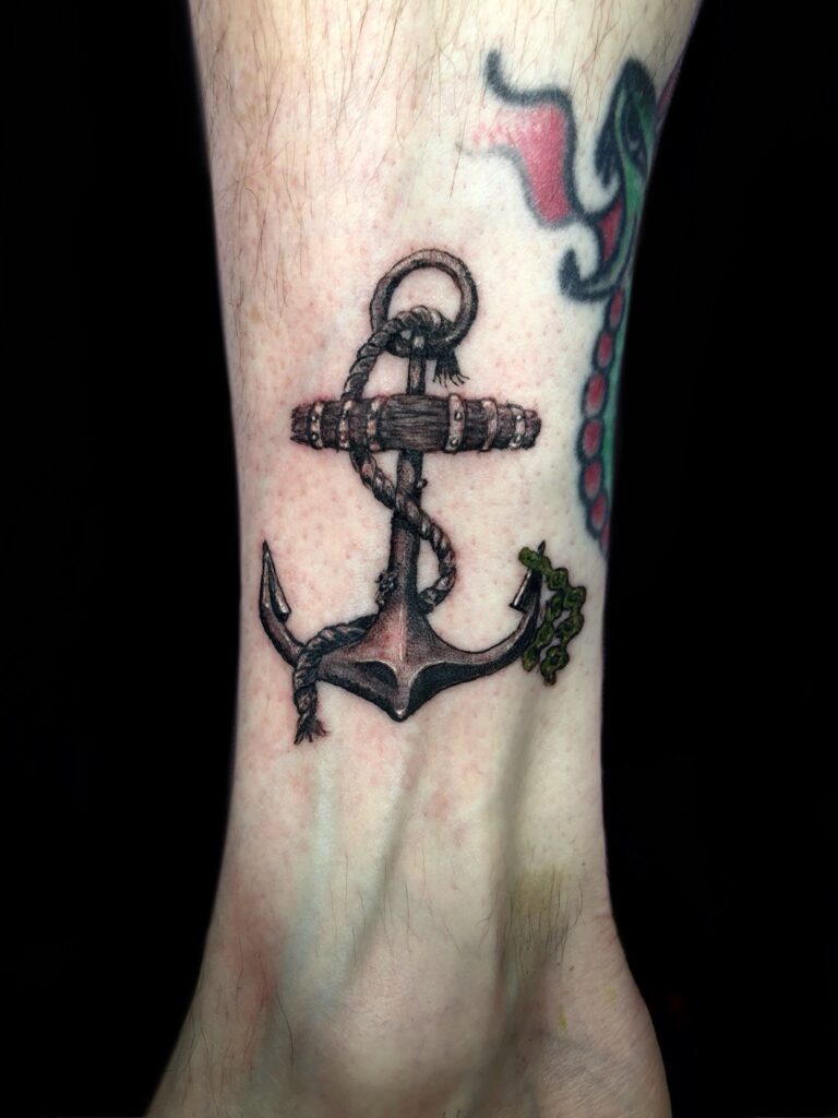 Detailed black anchor and rope tattoo with small green accents on a person’s lower leg in Roatan, Honduras.