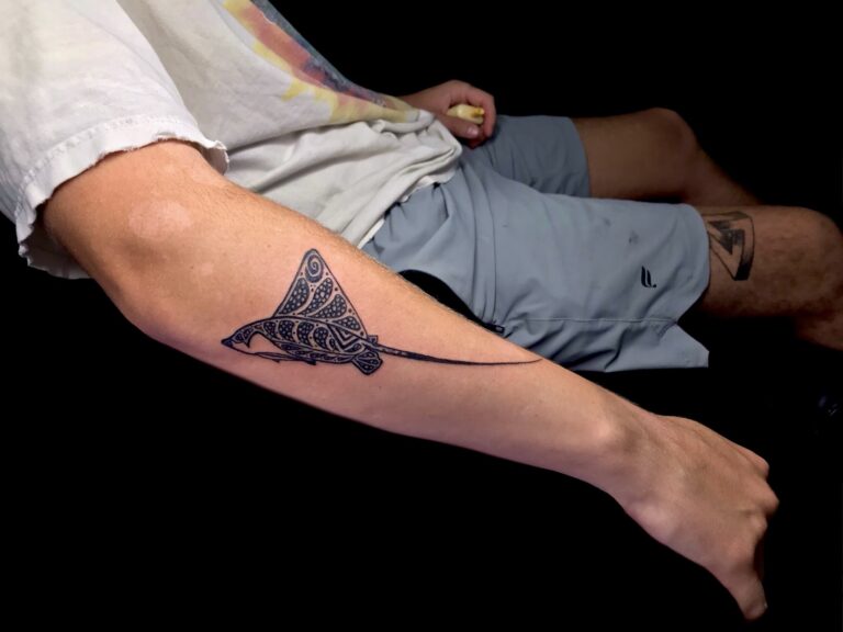 A seated person in shorts displays a detailed black tribal-style tattoo on the forearm in Roatan, Honduras.