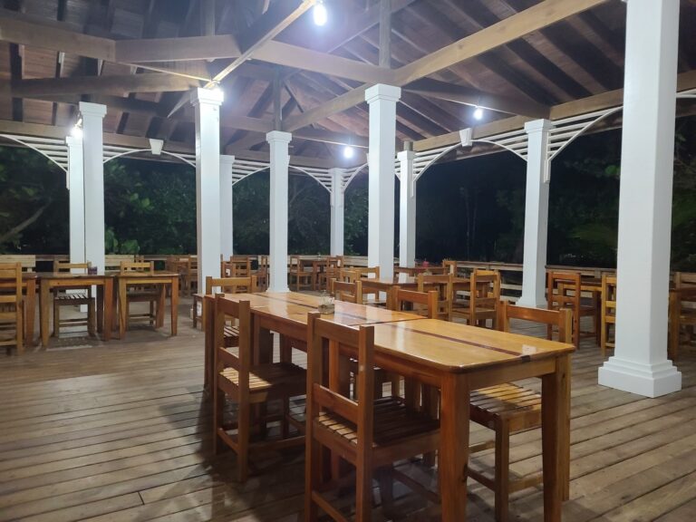 Open-air wooden pavilion in Roatan, Honduras, featuring white columns and a high peaked roof structure illuminated at night.