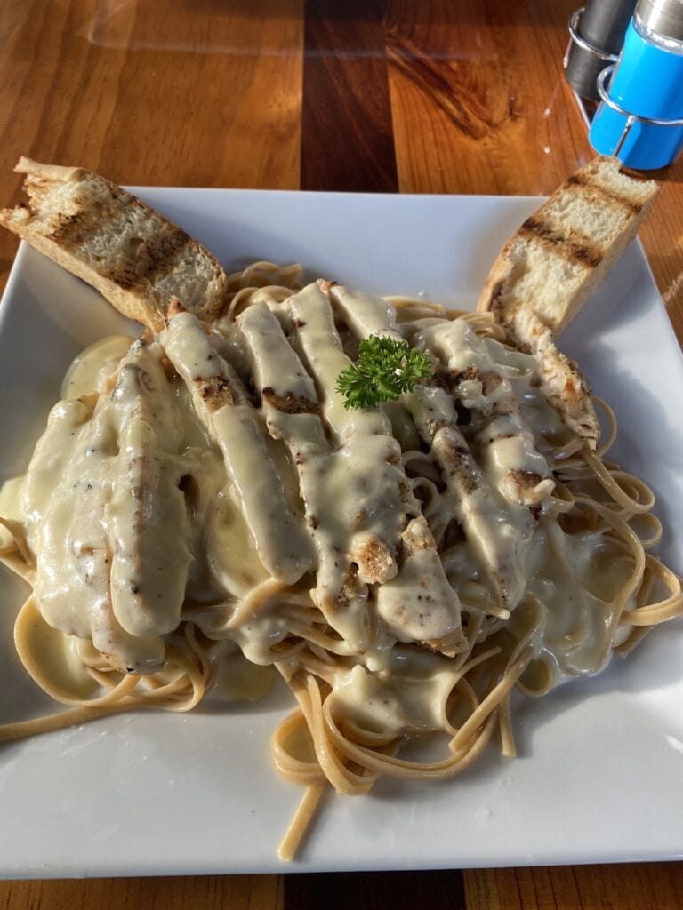 Creamy Alfredo pasta with grilled chicken served with toasted bread on a white plate set on a wooden table in Roatan Honduras.
