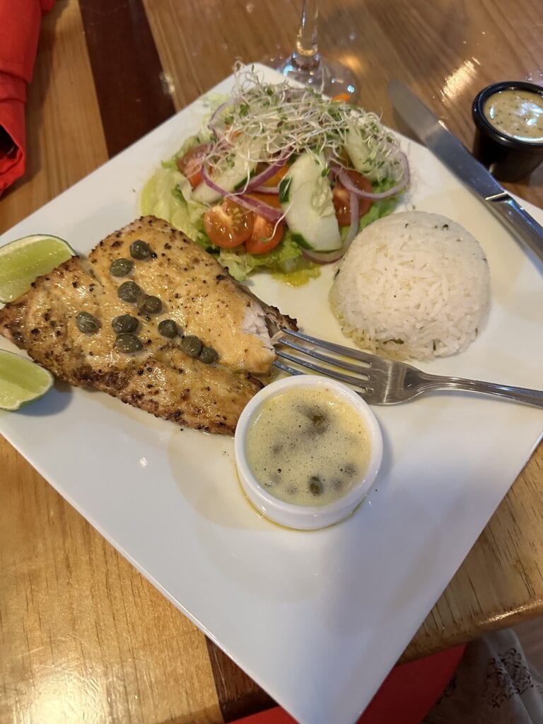 A plate of grilled fish topped with capers, served with white rice, a fresh salad with cherry tomatoes, cucumber, and sprouts, and lime wedges on a wooden table in Roatan, Honduras.