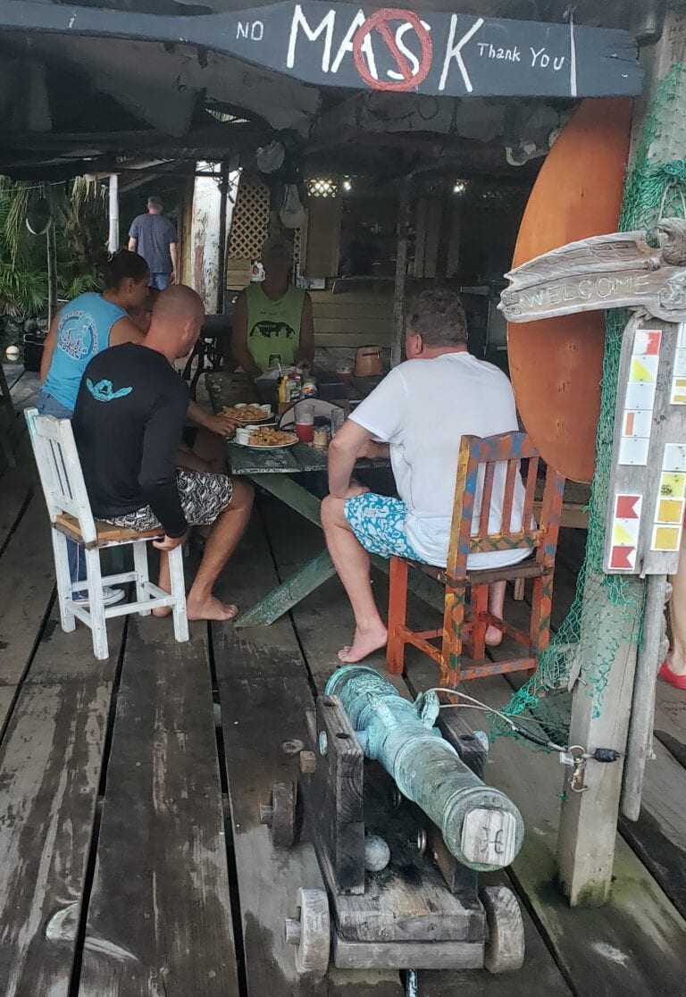 A group of four people seated at a rustic wooden table inside a casual restaurant with tropical decor on Roatan, Honduras, enjoying a meal together under a partially shaded roof.