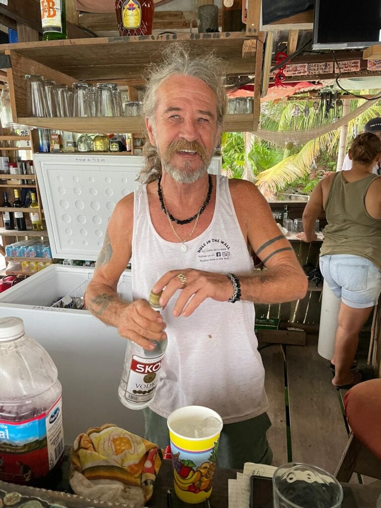 A cheerful man with long gray hair and a beard, holding a bottle of SKOL vodka inside a rustic bar in Roatan, Honduras, with tropical greenery visible through the window in the background.