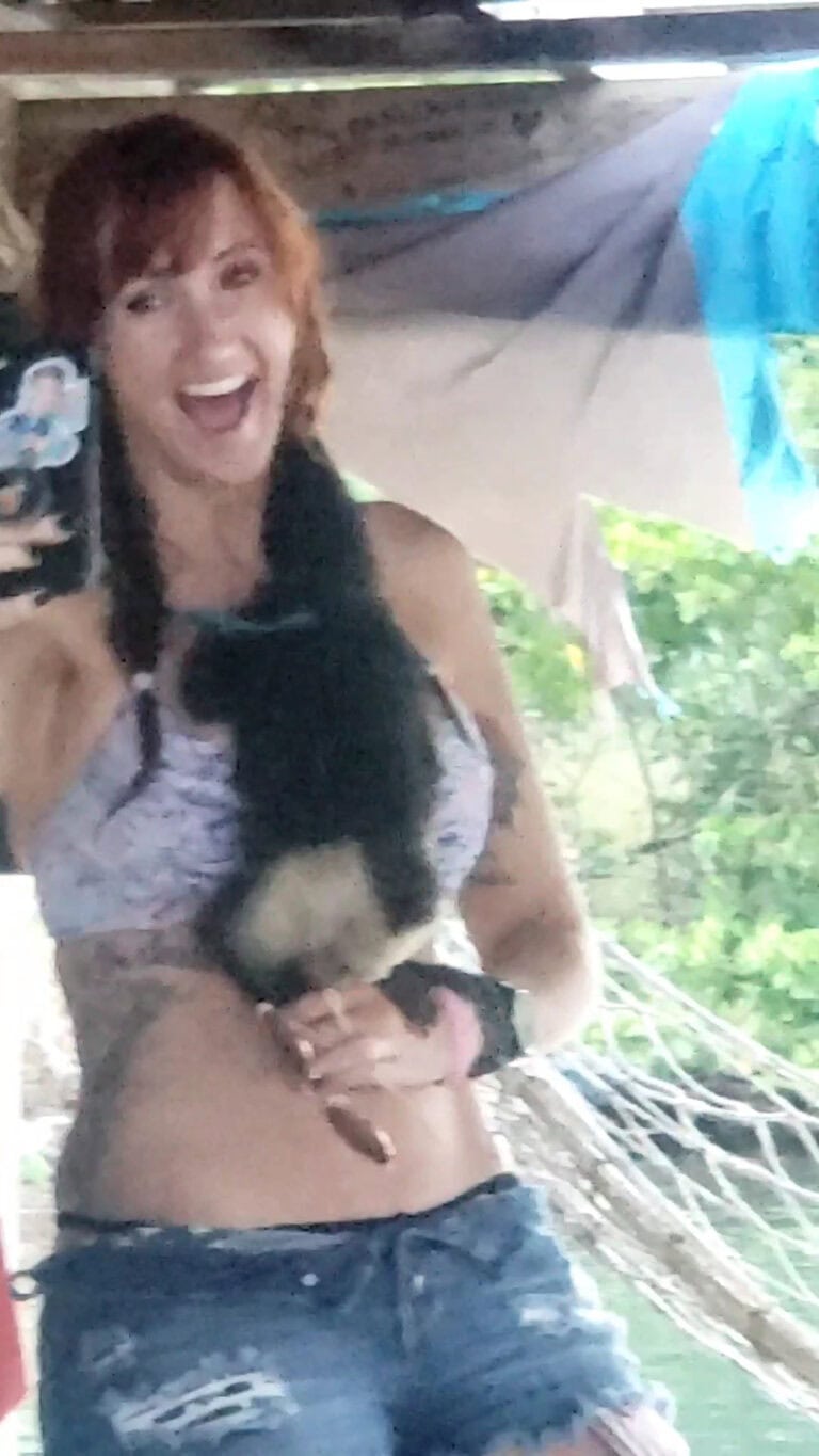A woman with reddish-brown hair and a black tank top holding a small puppy under a shaded outdoor structure in Roatan, Honduras.