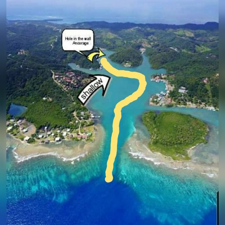 Aerial view of Roatan, Honduras, showing a winding shallow channel leading to an anchorage with a hole in the wall formation in the water and green hilly terrain surrounding the area.