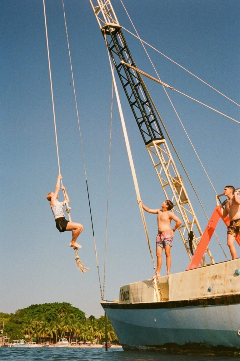 Children and adults enjoying a zip line adventure on Roatan, Bay Islands, Honduras, with lush green trees and a marina visible in the background under a clear blue sky.