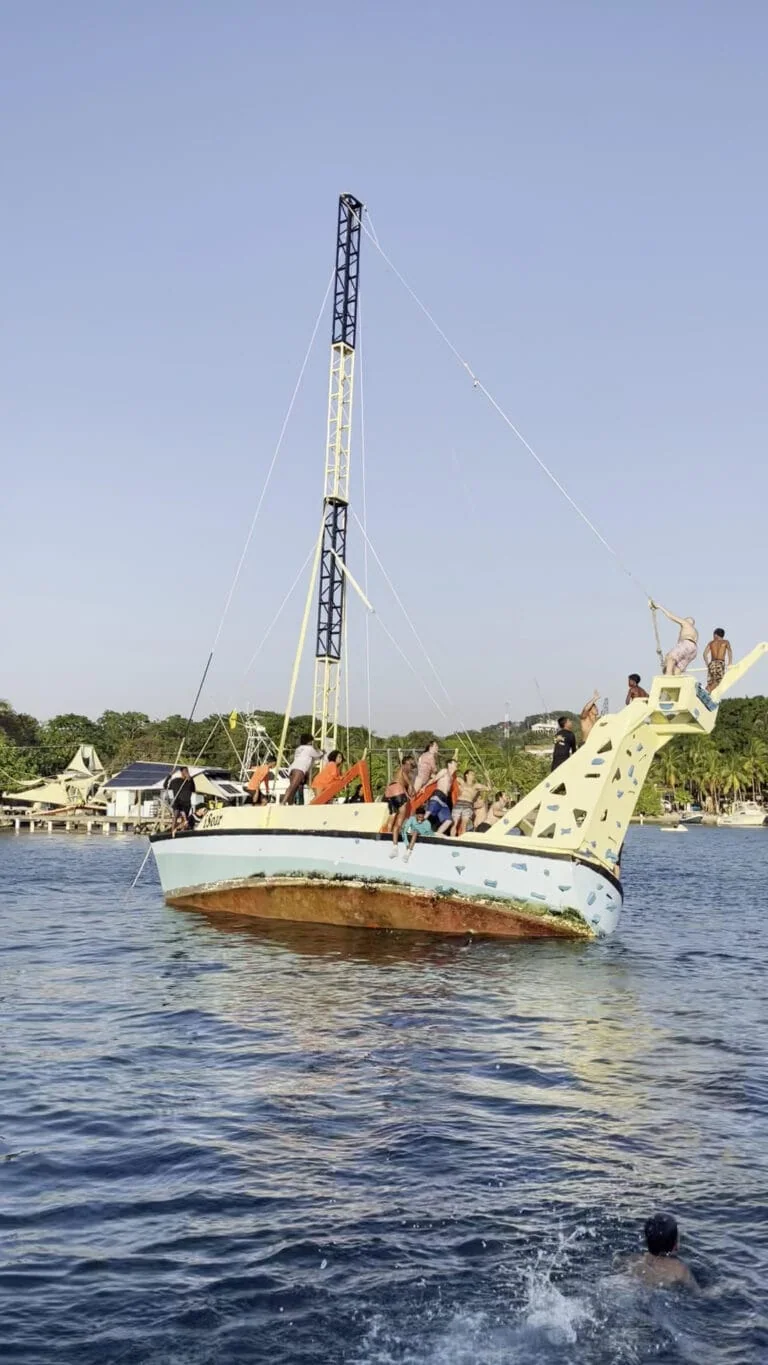 People enjoying a boat trip on a large whale-shaped vessel with a tall mast, anchored in the turquoise waters of Roatan, Honduras, with a coastal village and lush green trees in the background.