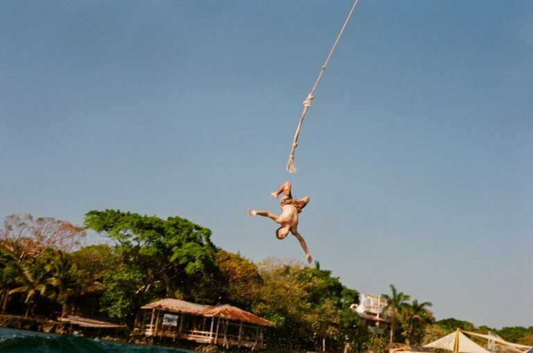 A person is mid-air on a rope swing over the water in Roatan, Honduras, with a lush green shoreline and thatched-roof huts in the background.