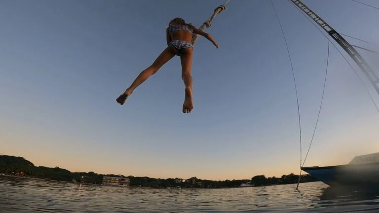 A young girl in colorful swimsuit swings high above the water on Roatan, Honduras, during sunset with the sky turning soft shades of orange and purple, and boats and shoreline visible in the background.