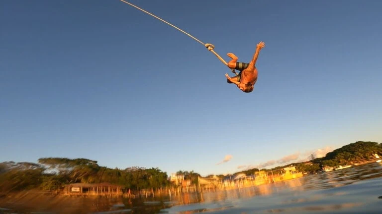 A man performing a high dive from a rope swing into the water with a backdrop of lush greenery and colorful buildings on Roatan, Honduras during sunset.