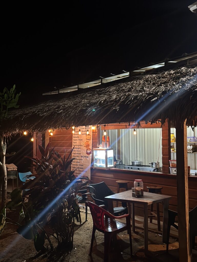 A warmly lit open-air restaurant patio in Roatan, Honduras, features a thatched roof, wooden bar counter, dining tables, chairs, tropical plants, and a wall menu.