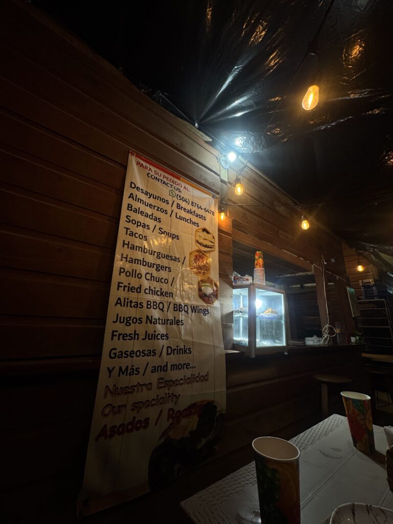 A bilingual food menu hangs beside a warmly lit wooden dining area in Roatan, Honduras, listing breakfasts, lunches, tacos, hamburgers, fried chicken, barbecue wings, fresh juices, and other specialties.