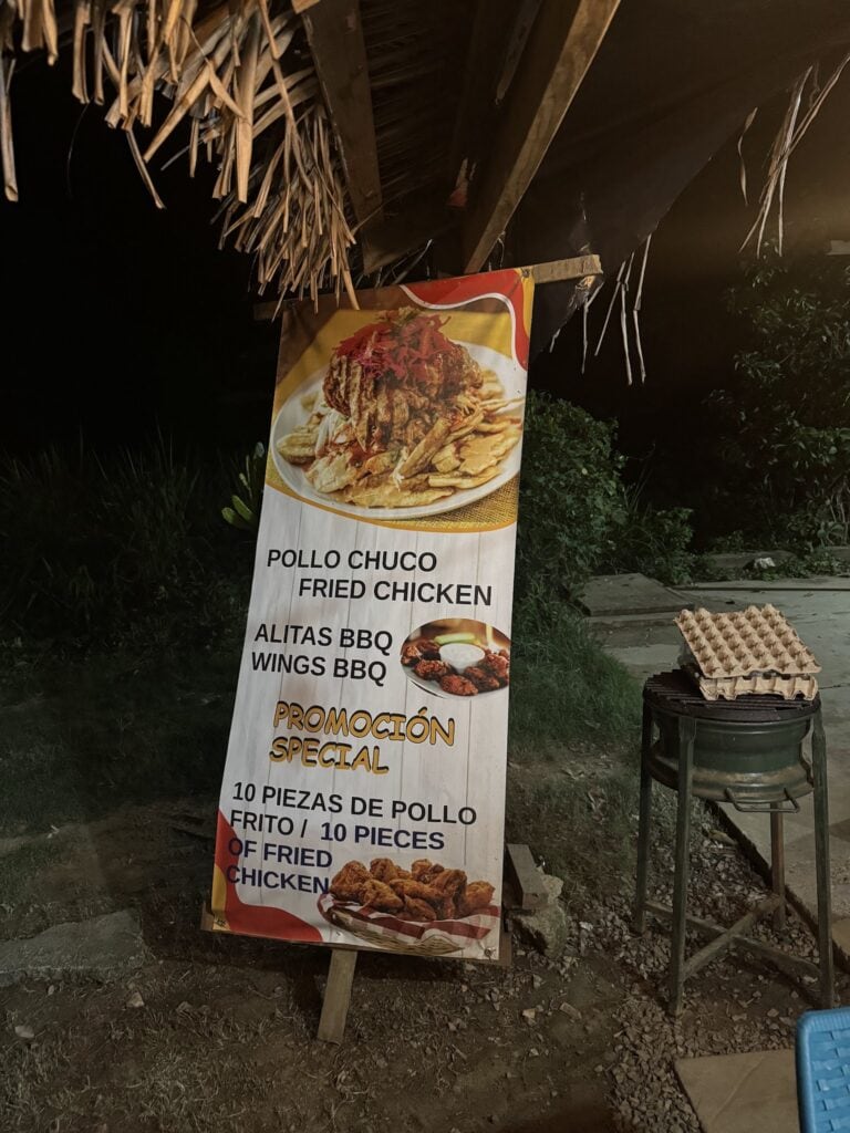 Promotional fried chicken sign advertising pollo chuco, BBQ wings, and a ten-piece special beneath a thatched roadside shelter in Roatan, Honduras.
