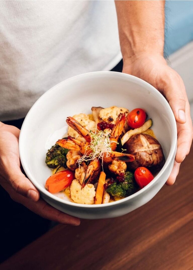 A bowl of grilled shrimp, roasted vegetables, tomatoes, broccoli, mushrooms, and sprouts is held over a wooden table in Roatan, Honduras.