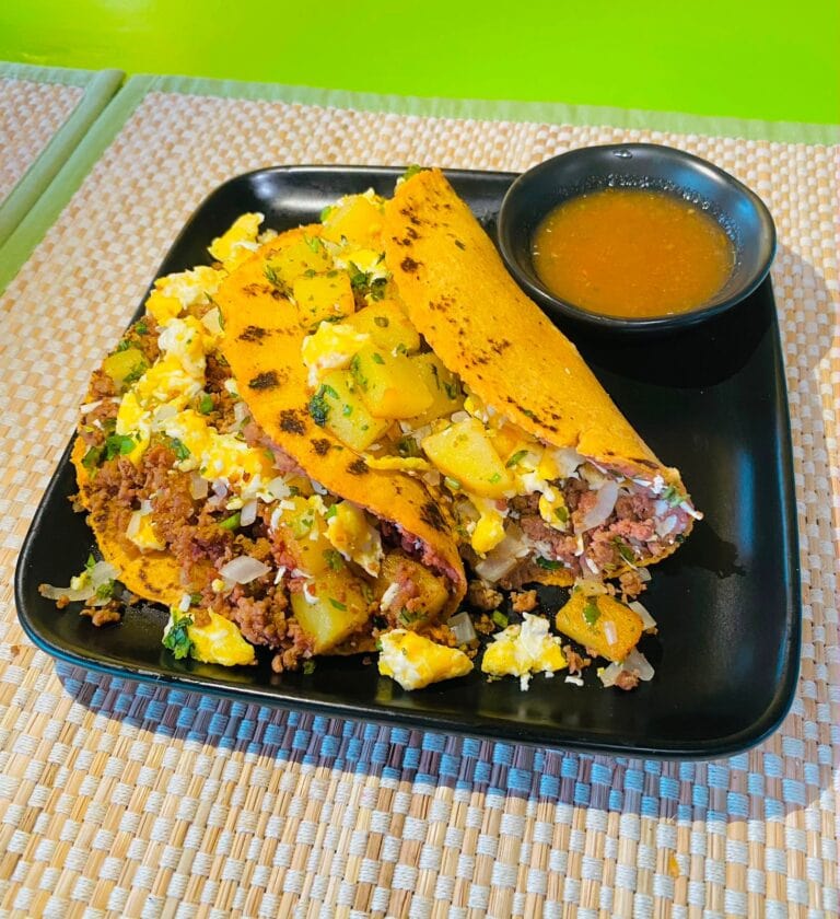 A black plate holds two golden stuffed tacos filled with scrambled eggs, seasoned ground meat, potatoes, vegetables, and cheese beside a bowl of orange dipping sauce in Roatan, Honduras.