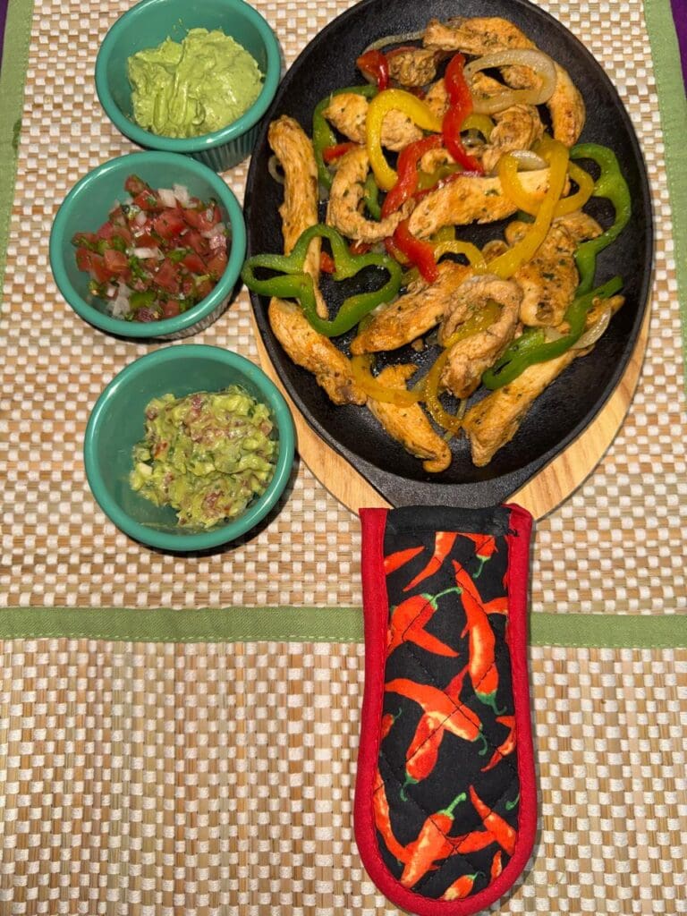 A black skillet holds grilled chicken strips with red, yellow, and green peppers beside three bowls of guacamole and fresh salsa on a woven placemat in Roatan, Honduras.