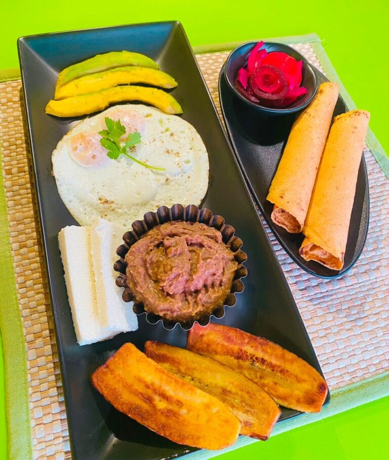 A Honduran breakfast platter features fried eggs, avocado slices, plantains, tortillas, cheese, refried beans, and pickled vegetables in Roatan, Honduras.