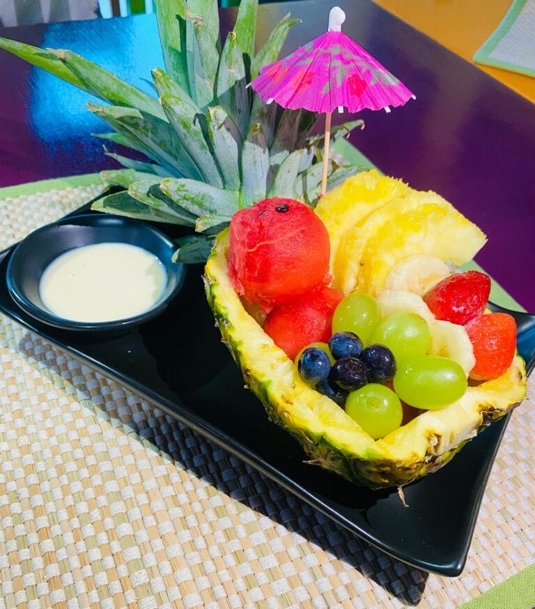 A pineapple fruit bowl filled with watermelon, pineapple, strawberries, grapes, blueberries, and dipping sauce sits on a black plate in Roatan, Honduras.