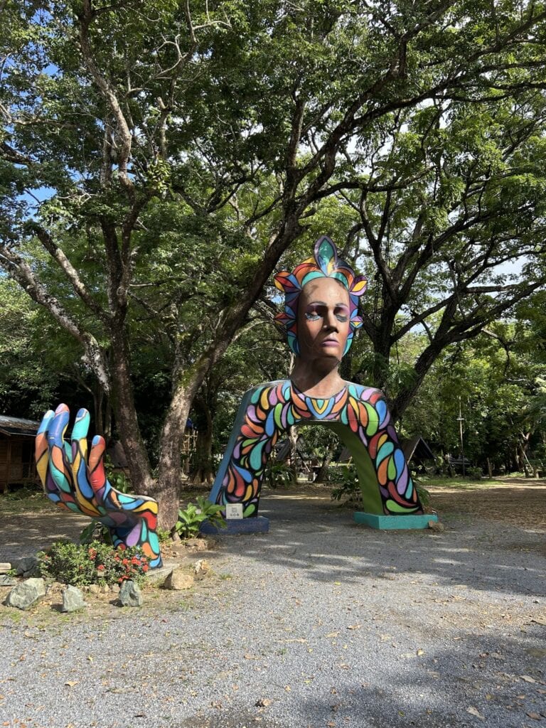 A colorful, painted sculpture of a woman's face with intricate, vibrant patterns and headpieces stands outdoors among trees in Roatan, Honduras.