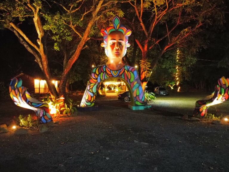 A illuminated colorful sculpture of a woman with a large headpiece and stylized features stands in a garden in Roatan, Honduras, surrounded by trees, benches, and parked cars at night.