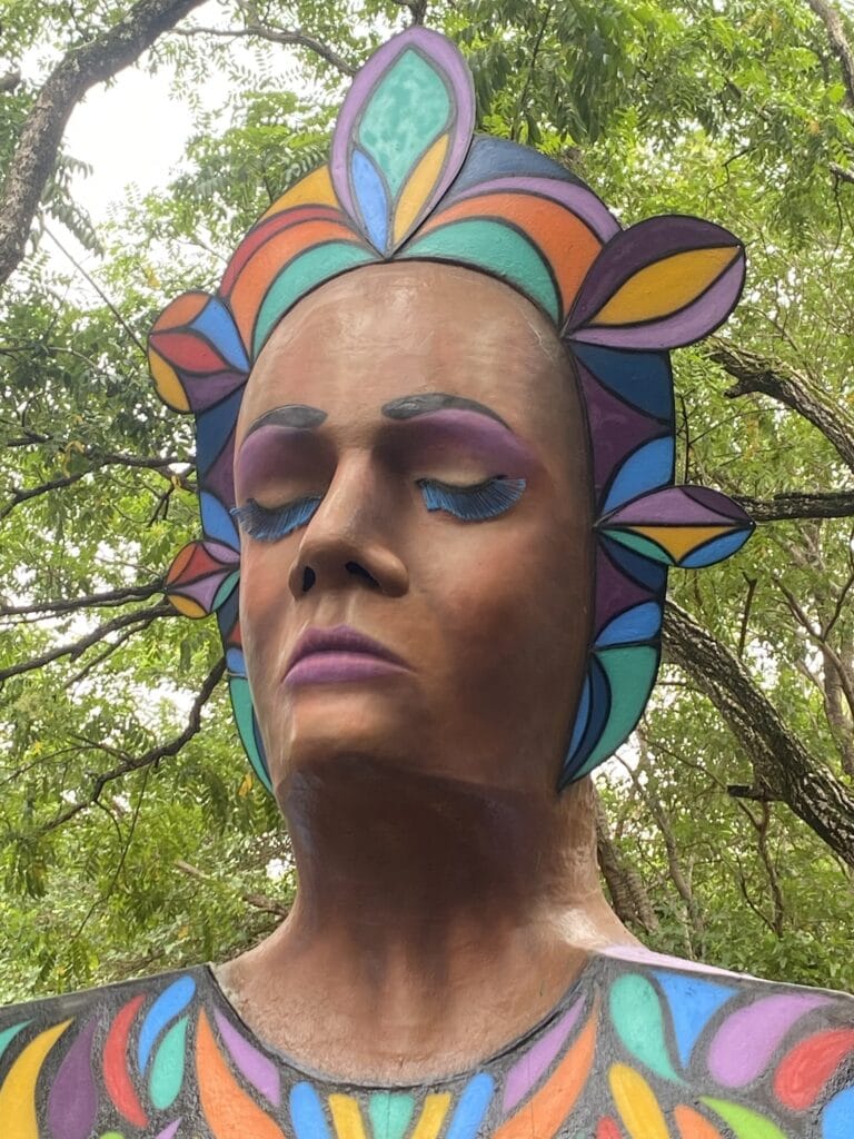 A painted mural of a woman's face with closed eyes and colorful, stylized leaves or petals surrounding her head, set outdoors among green trees in Roatan, Honduras.