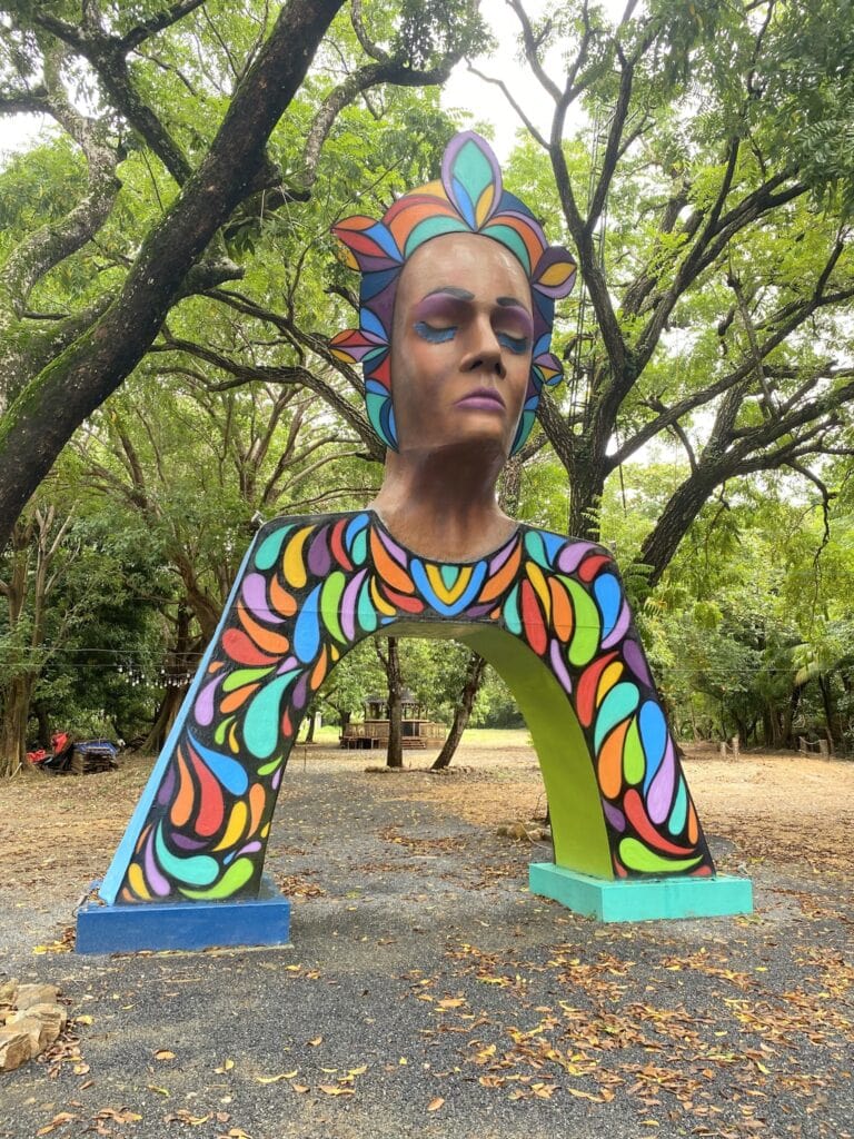 A colorful painted sculpture of a woman's face with closed eyes and vibrant, patterned hair and clothing, located outdoors among trees in Roatan, Honduras.