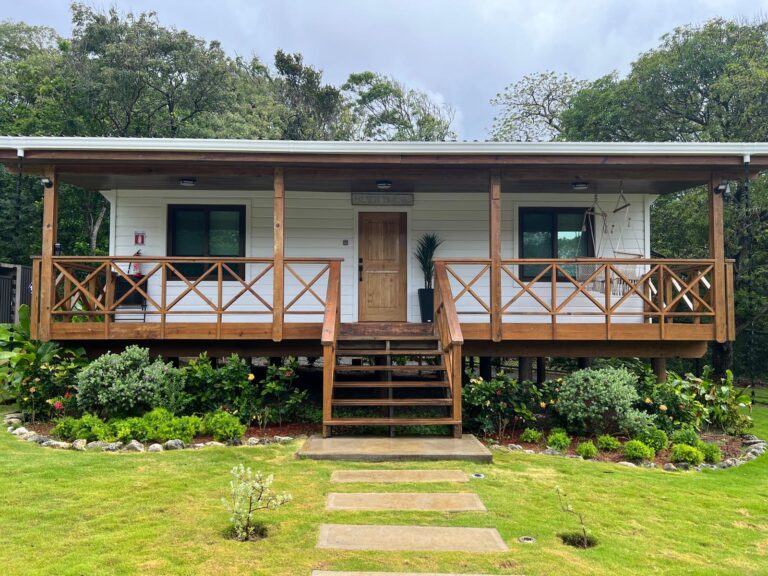 A single-story wooden bungalow with a covered veranda, front steps, and landscaped garden in Roatan, Honduras.