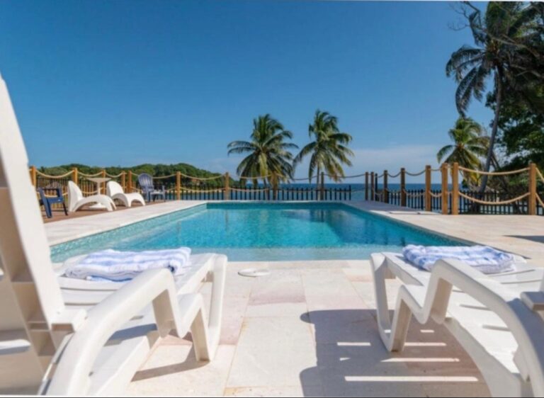 Turquoise resort pool with white lounge chairs, palm trees, and Caribbean Sea views in Roatan, Honduras.