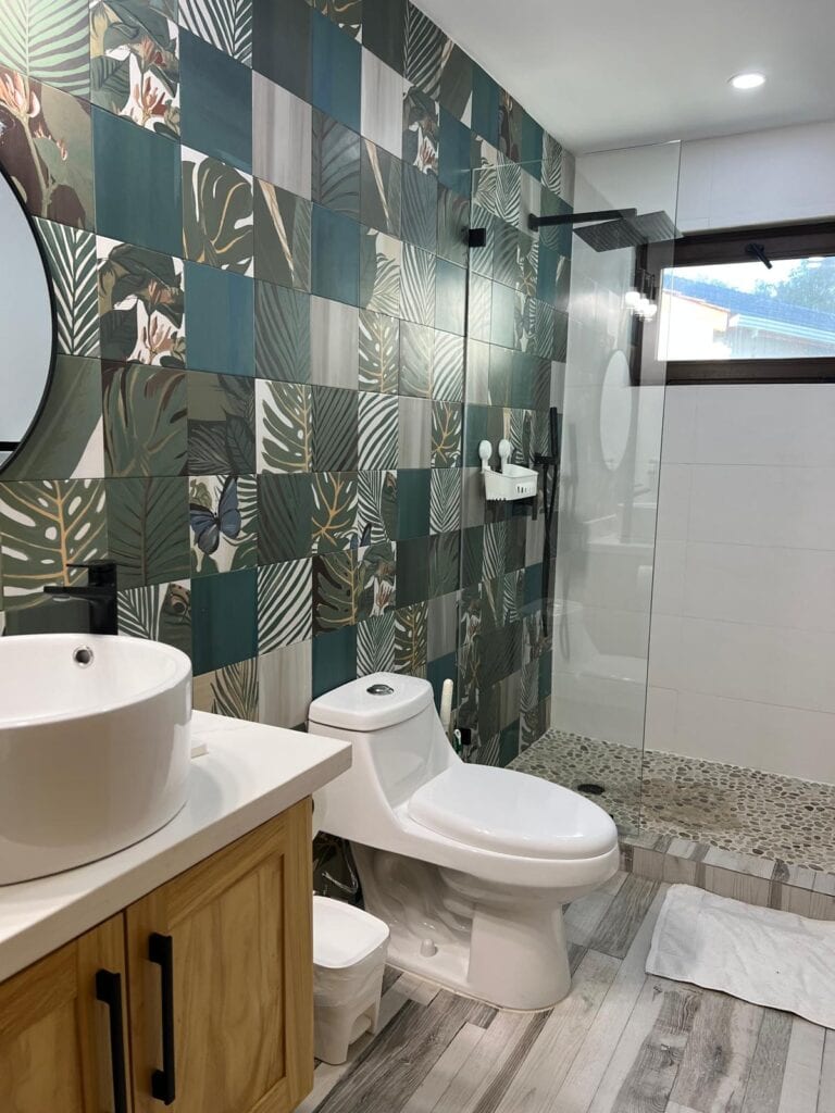 Modern bathroom in Roatan, Honduras, featuring a patterned botanical tile wall, vessel sink, toilet, and glass-enclosed shower.