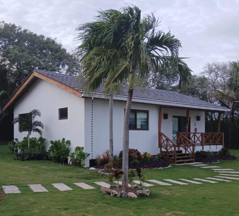 A modern tropical cottage with a tiled roof, wooden porch, landscaped lawn, and palm trees in Roatan, Honduras.
