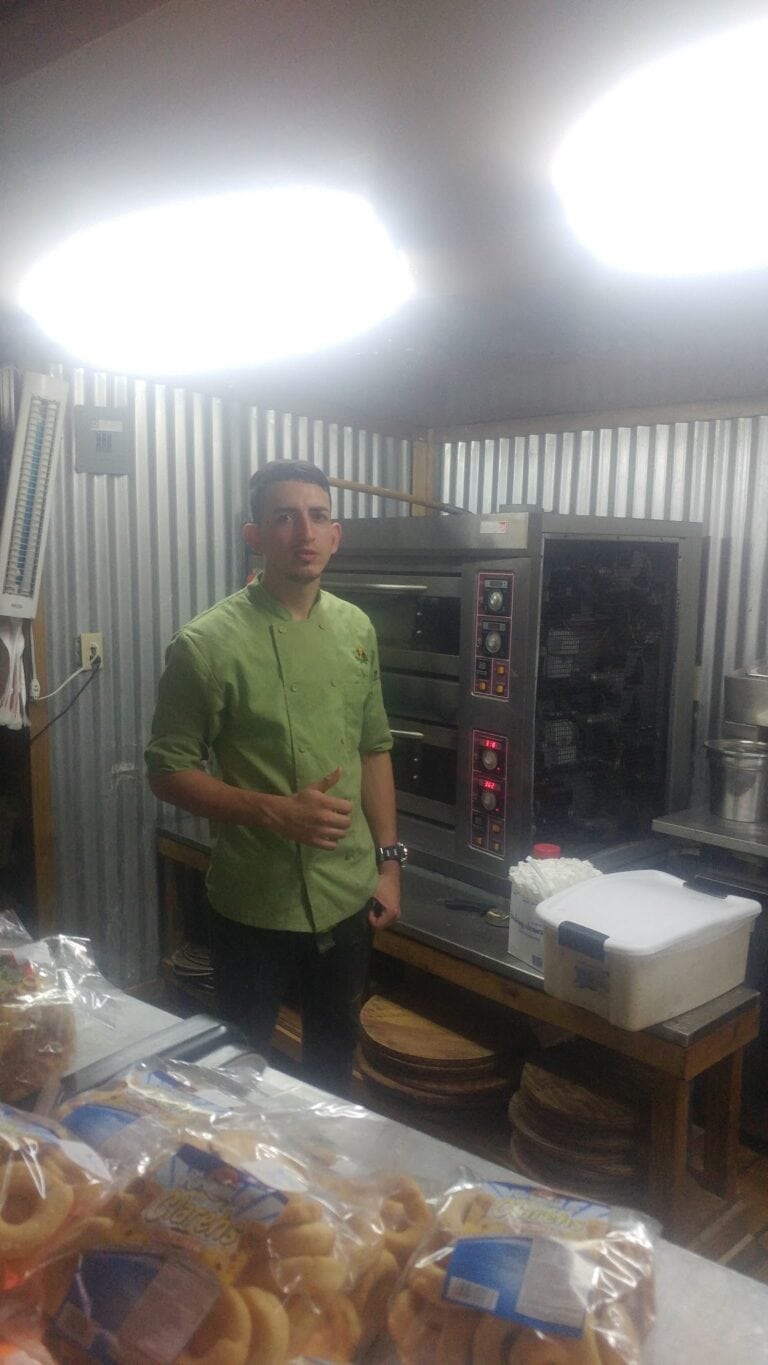 A young man wearing a green chef's jacket gives a thumbs-up in a commercial kitchen with industrial ovens and baking supplies, located on Roatan in the Bay Islands of Honduras.