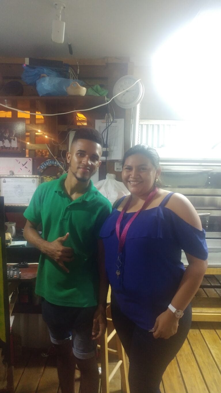 A young man and a woman stand together inside a restaurant on Roatan, Bay Islands, Honduras, smiling at the camera with a busy kitchen background behind them.