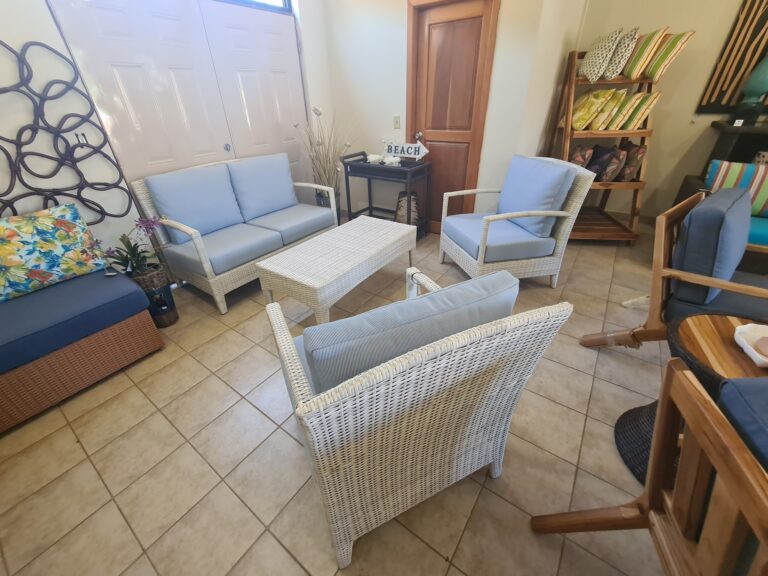 A cozy indoor seating area on Roatan, Honduras, features wicker and wooden chairs with light blue and patterned cushions, a small black table with a "Beach" sign, decorative pillows, and a wooden shelving unit with colorful pillows against a beige wall.