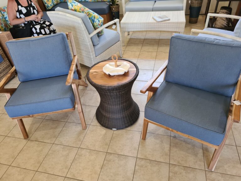 A seating area at a furniture store in Roatan, Honduras, features wicker and wooden chairs with blue cushions, a white wicker coffee table with magazines, and decorative pillows with floral patterns, set on a tiled floor.