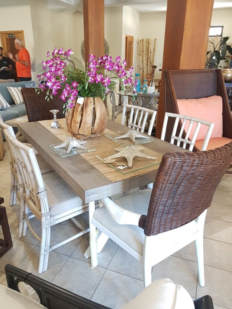 A dining area in Roatan, Honduras, features a wooden table with a decorative flower arrangement in a rustic wooden vase, surrounded by wicker and white chairs, with tropical decor including starfish and seashells on the table.